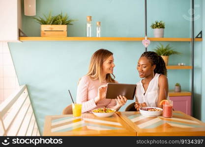 Two pretty young black and caucasian woman having good time, drinking fresh juices and having healthy breakfast in the cafe