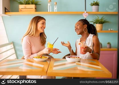 Two pretty young black and caucasian woman having good time, drinking fresh juices and having healthy breakfast in the cafe