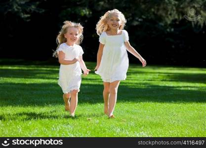 Two pretty young blond girls wearing white dresses and having fun running through a green park in summer sunshine