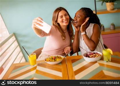 Two pretty young women, caucasian and black one, taking selfie with mobile phone in the cafe