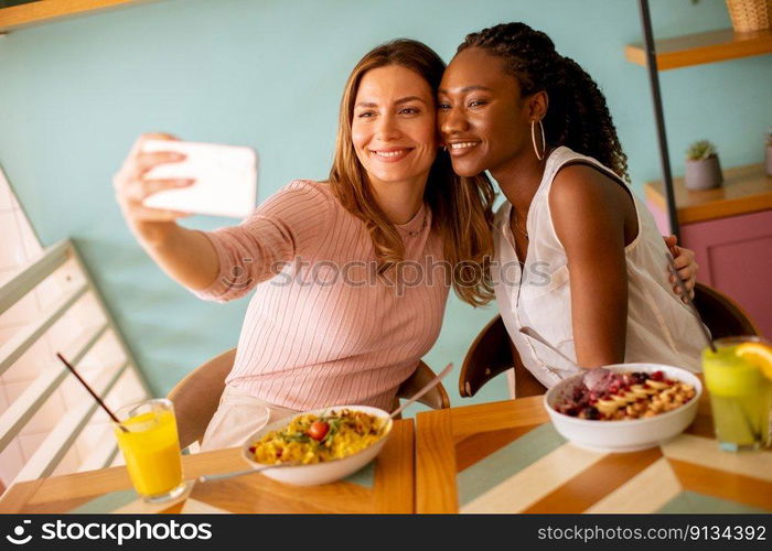 Two pretty young women, caucasian and black one, taking selfie with mobile phone in the cafe