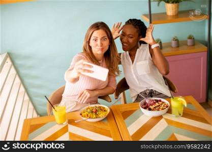 Two pretty young women, caucasian and black one, taking selfie with mobile phone in the cafe