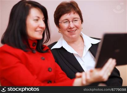 Two professional business women sharing a tablet computer smile in amusement at something on the screen with focus to an attractive middle-aged woman in glasses