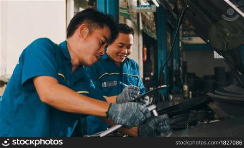 Two professional car mechanic screwing details of car engine on lifted automobile at repair service station. Skillful Asian guy in uniform fixing car at garage night. Car service concept.