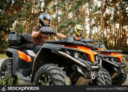 Two quad bike riders in helmets and equipment, side view, closeup, summer forest on background. Male quadbike drivers, atv riding, extreme sport. Two quad bike riders in helmets closeup, side view