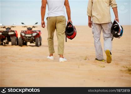 Two racers hold helmets near atvs, freedom riding in desert. Male persons on quad bikes, sandy race, dune safari in hot sunny day, 4x4 extreme adventure, quad-biking. Two racers hold helmets near atvs in desert