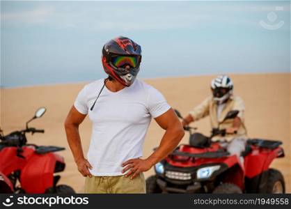 Two racers in helmets poses near atvs, freedom riding in desert. Male persons on quad bikes, sandy race, dune safari in hot sunny day, 4x4 extreme adventure, quad-biking. Two racers in helmets poses near atvs in desert