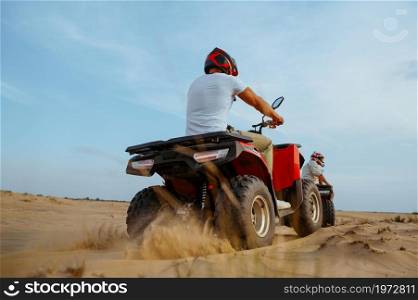 Two racers in helmets ride on atv, action view, freedom riding in desert. Male persons on quad bikes, sandy race, dune safari in hot sunny day, 4x4 extreme adventure, quad-biking. Two racers in helmets ride on atv, action view