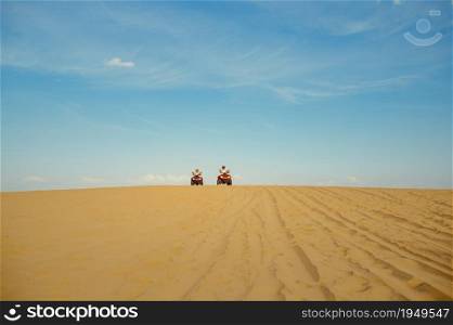 Two racers in helmets riding on atv in desert sands, afar view. Male persons on quad bikes, sandy race, dune safari in hot sunny day, 4x4 extreme adventure, quad-biking. Two racers riding on atv in desert, afar view