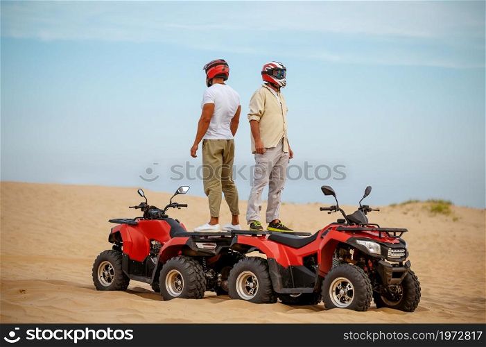 Two racers in helmets stand back to back on atvs, freedom riding in desert. Male persons on quad bikes, sandy race, dune safari in hot sunny day, 4x4 extreme adventure, quad-biking. Two racers in helmets stand back to back on atvs