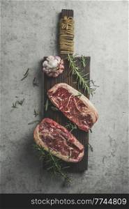 Two raw marbled rib-eye steak on cutting board with rosemary, garlic and green pepper on grey concrete kitchen table. Barbecue preparation with fresh meat. Top view.