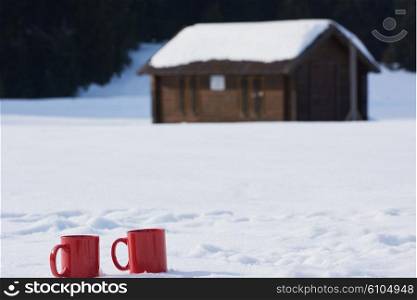 two red coups of hot tea drink in snow at beautiful winter sunny day scene with wooden house in background in forest