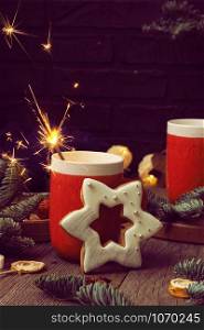 Two red mugs with coffee and marshmallows, a burning sparkler, star-shaped gingerbread and fir branches on a wooden background.