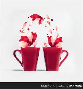 Two red mugs with red white flying flowers and petals of Amaryllis flowers standing on white background. Creative couples concept. Romantic holiday layout.