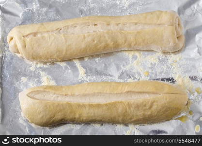 Two rolled up batons of Italian-style bread dough, glazed with egg-wash, slashed and left to rise on a baking sheet