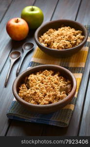 Two rustic bowls of baked apple crumble or crisp on kitchen towel, wooden spoons and fresh apples on the side, photographed on dark wood with natural light (Selective Focus, Focus one third into the first crumble)