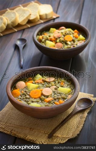Two rustic bowls of lentil soup made with potato, carrot, onion and sausage slices, with slices of baguette bread in the back, photographed on dark wood with natural light (Selective Focus, Focus on the middle of the first soup)