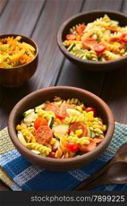 Two rustic bowls of pasta salad made of tricolor fusilli, sweet corn, cucumber, cherry tomato and sausage, photographed with natural light (Selective Focus, Focus one third into the first salad)