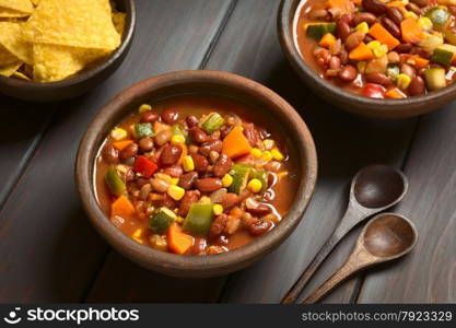 Two rustic bowls of vegetarian chili dish made with kidney bean, carrot, zucchini, bell pepper, sweet corn, tomato, onion, garlic, with tortilla chips on the side, photographed with natural light (Selective Focus, Focus in the middle of the first dish)