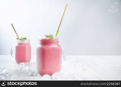 Two rustic jars with fresh blended smoothie from yogur, berries and ice isolated on wooden table and white background surrounded with melted ice, golden straw inside Healthy summer drink