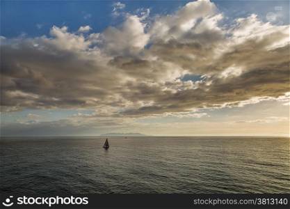 Two sailing boats pass a silhouetted island of Capri with blue skies and clouds above