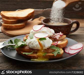 two sandwiches on a toasted white slice of bread with poached eggs, green leaves of arugula and radish in a round plate, behind a cup of coffee, morning breakfast