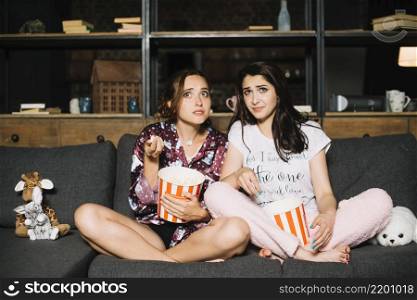 two scared women sitting sofa with popcorn watching television