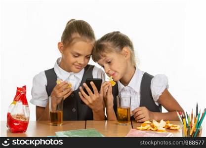 Two schoolgirls at a break look at the screen of a smartphone, and eat an orange and drink juice