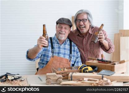 Two senior carpenter men Asian and Caucasian celebrating together with couple bottle of beer after finished their wood work at workshop. Working happily in retirement conceptnt concept