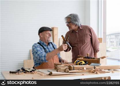 Two senior carpenter men Asian and Caucasian holding and cheers bottle of beer together after finished their wood work at workshop. Working happily in retirement concept