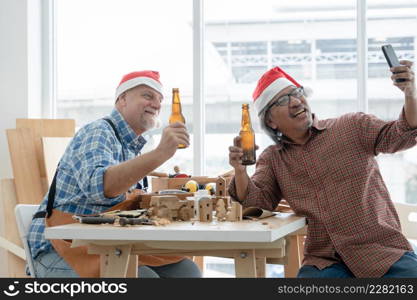 Two senior carpenter men Caucasian and asian with beard wearing santa hat celebrating with beer bottles and selfie with mobile phone after finished on working wooden at workshop on Christmas holiday