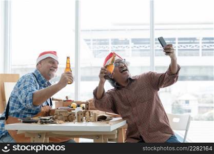 Two senior carpenter men Caucasian and asian with beard wearing santa hat celebrating with beer bottles and selfie with mobile phone after finished on working wooden at workshop on Christmas holiday