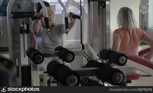 Two senior women working out on exercisers in fitness centre. One person using chest press machine and the other pull back machine