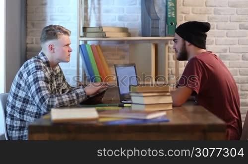 Two serious male students in library surrounded by books using digital devices and studying for exams. Young teenagers sitting face to face at the table and learning. Hipster with beard and hat working with laptop, his friend using digital tablet.