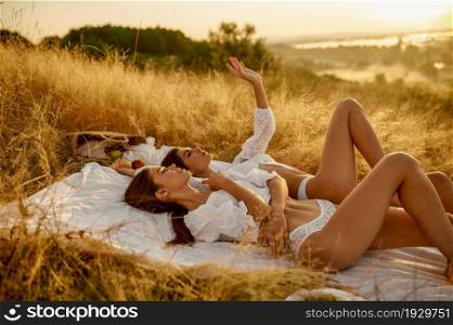 Two sexy women in lingerie lying on blanket in the field. Female persons with slim body in underwear leisures on meadow, relaxation on sunset, feeling of freedom. Two women in lingerie lying on blanket in field