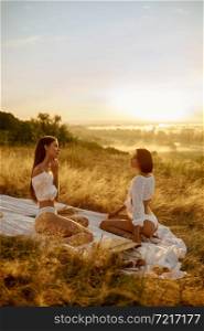 Two sexy women in lingerie sitting on blanket in the field. Female persons with slim body in underwear leisures on meadow, relaxation on sunset, feeling of freedom. Two sexy women in lingerie on blanket in the field