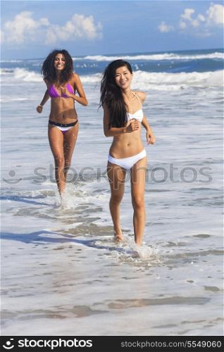 Two sexy young bikini wearing women or girls running through the surf on a deserted tropical beach with a blue sky, one girl is Chinese Asian the other mixed race Hispanic