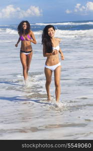 Two sexy young bikini wearing women or girls running through the surf on a deserted tropical beach with a blue sky, one girl is Chinese Asian the other mixed race Hispanic