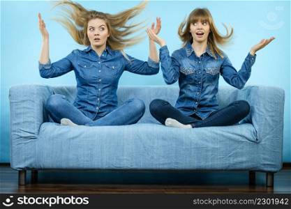 Two shocked women wearing jeans shirts having windblown blonde hair. Blue background.. Two shocked women with windblown hair