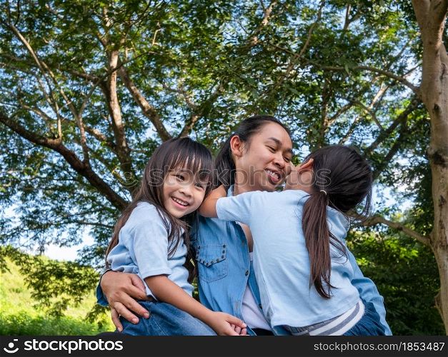 Two sibling little girls with mom laughing and hugging each other on warm and sunny summer day in the garden. Young girls with her mother spending day in park.