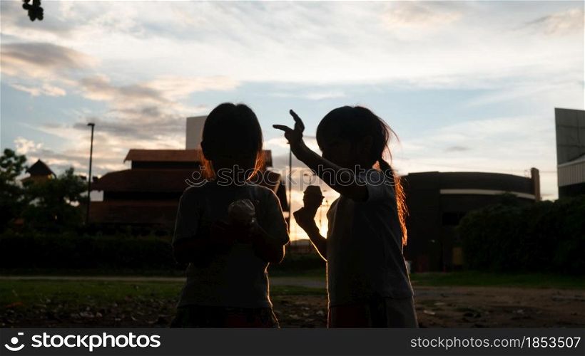 Two sibling sisters eating sweet tasty ice cream outdoors at sunset. Two happy young kid enjoy dessert during vacation holiday in the park.