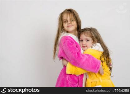 Two sister girls with wet hair standing in the bath robes on a light background. Girls in gowns hug after swimming