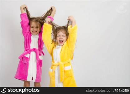 Two sister girls with wet hair standing in the bath robes on a light background. Two girls in the bath robes raised their wet hair