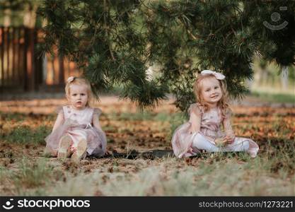 two sisters in identical dresses sit under a tree on the ground and smile. Happy childhood. Little girls having fun. two sisters in identical dresses sit under a tree on the ground and smile. Happy childhood. Little girls having fun.