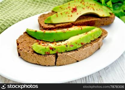 Two slices of rye bread with slices of avocado and pepper on a plate, green cloth, parsley on the background light wooden boards