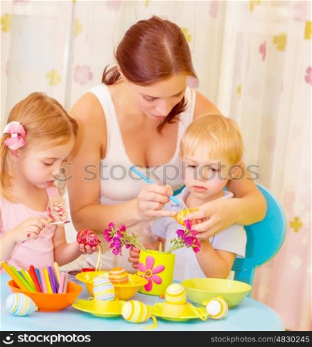 Two small cute children with beautiful mother paint Easter eggs at home, mom teaching babies decorate traditional eastertime symbol