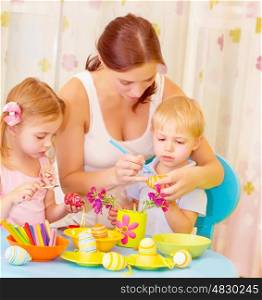 Two small cute children with beautiful mother paint Easter eggs at home, mom teaching babies decorate traditional eastertime symbol