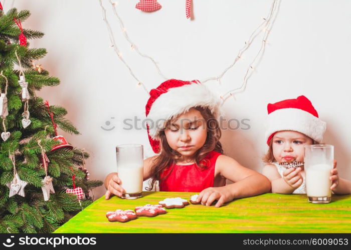 Two small Santas are drinking milk and eating colorful gingerbreads. Two girls near Christmas tree