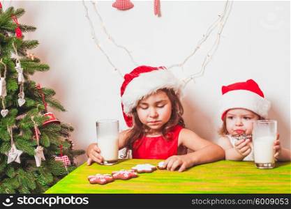 Two small Santas are drinking milk and eating colorful gingerbreads. Two girls near Christmas tree