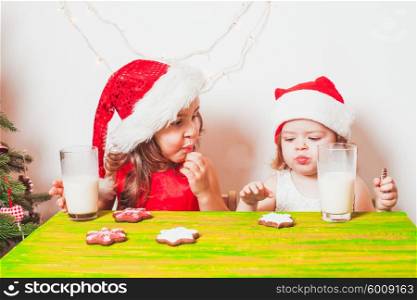 Two small Santas are drinking milk and eating colorful gingerbreads. Two girls near Christmas tree